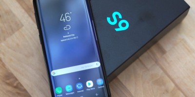 Galaxy S9 preto com a tela acionada, apoiado na caixa do aparelho, preta e com a inscrição "S9" em azul turquesa. Ambos estão dispostos sobre uma mesa de madeira.