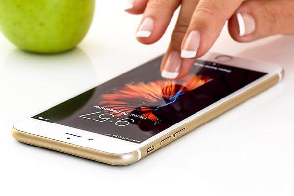 Mulher mexendo em um iPhone dourado, que está sobre a mesa, com uma maçã verde ao fundo. O papel de parede do celular é de fogos de artifício.