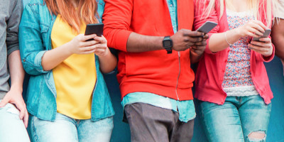 Foto de jovens encostados em uma parede azul. Cada um deles segura um celular nas mãos.