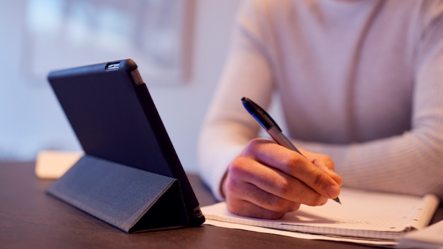 Foto de uma tablet em cima da mesa e uma mão segurando uma caneta em sua frente.
